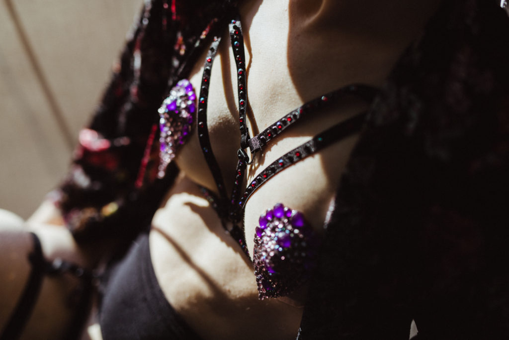 A close up of his purple bedazzled pasties sticking to his nipples and a harness that is bedazzled with red and black rhinestones. 