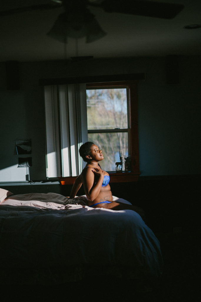 Kenni sits on the edge of a bed with a window in the background. Pictures are sticky tacked to the wall. A patch of sunlight enters and lights up Kenni in the otherwise shadowed surroundings. Their hand is on their chest and they close their eyes as they feel the sunlight.