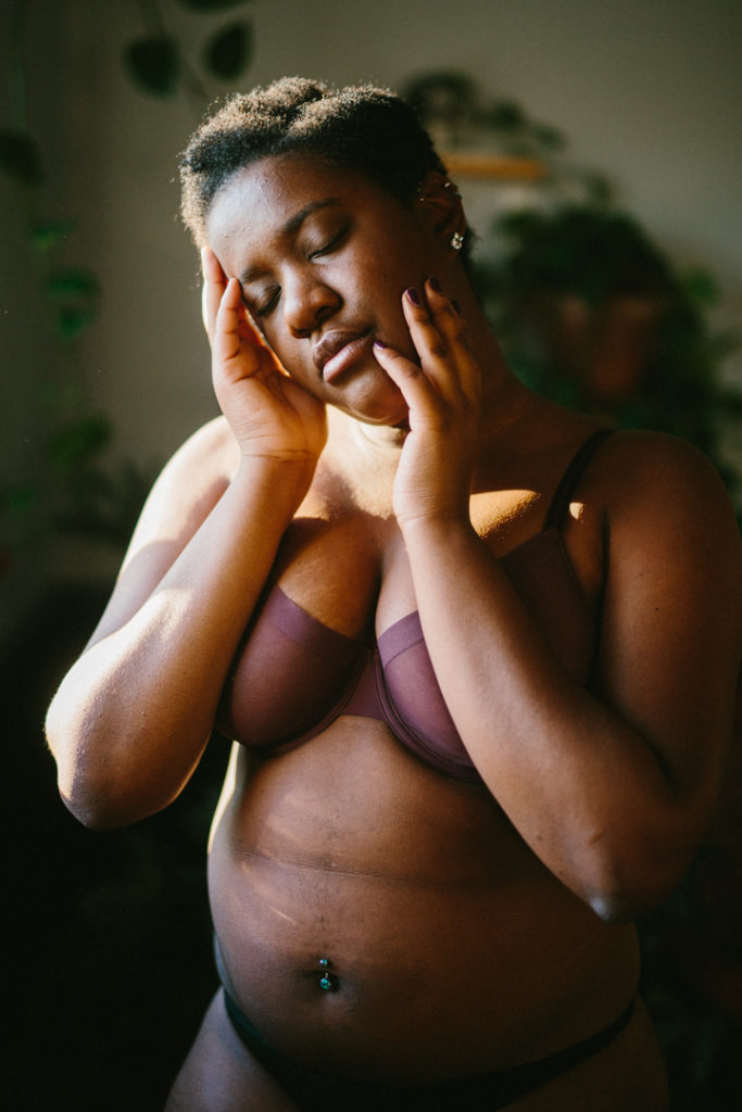 A waist up photograph of Kenni inside of the home studio. Light comes from a nearby widow, highlighting the features of their face. They raise one hand to their cheek and the other cradles their temple They're wearing a purple sheet bra and black panties. Green blurs are in the background, they're house plants. 