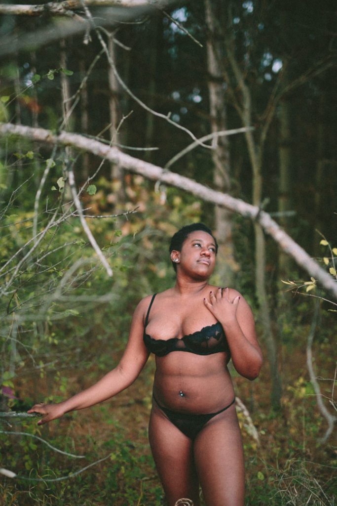 Kenni, a non-binary model from Chicago, stands center of the frame with their left hand on their chest and their right hand on a nearby branch. The frame has a big branch in is through the center, Kenni is positioned to be in the clearing. They wear a see through black bra and a black thong. Their ears and belly button are pierced. 