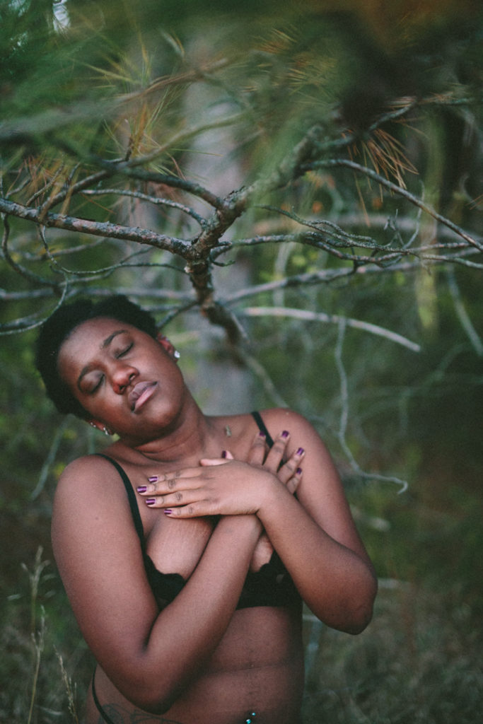 A cool temperature and true to color photograph of Kenni, a Black non-binary person who uses they/them pronouns. They have short black hair beautiful lips, their eyes are closed, and their arms are crossed as they gently embrace themself. Their head is tilted to the side and there are faint stretch marks on their breasts. A huge branch from a pine tree extends outward into the top of the frame, its green bristles fill it.