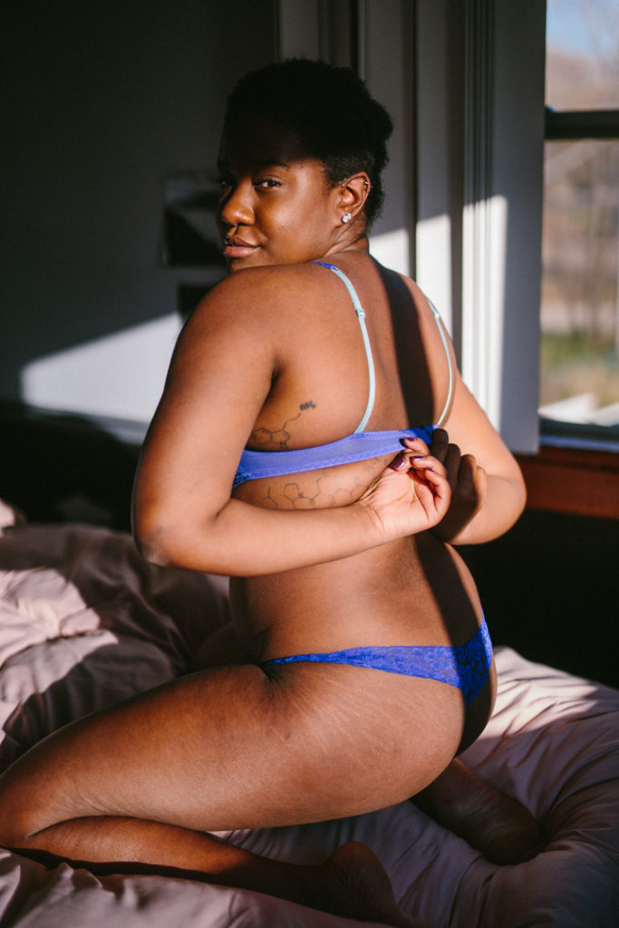 Kenni sits on their knees and unclasps their bra in a patch of sunlight on the bed. Their bra is vibrant blue, faint stretch marks line the side of their butt and thighs. They have a tattoo of a chemical compound sequence on their left upper side.